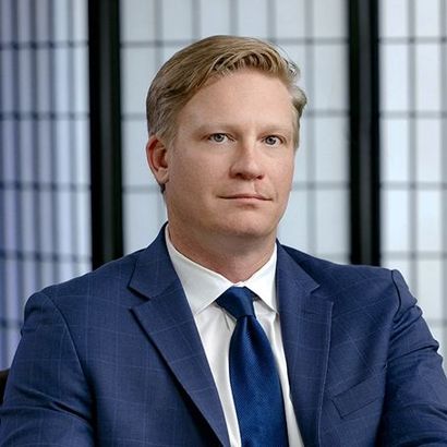 A person in a blue plaid suit and blue tie posing against a white grid background.