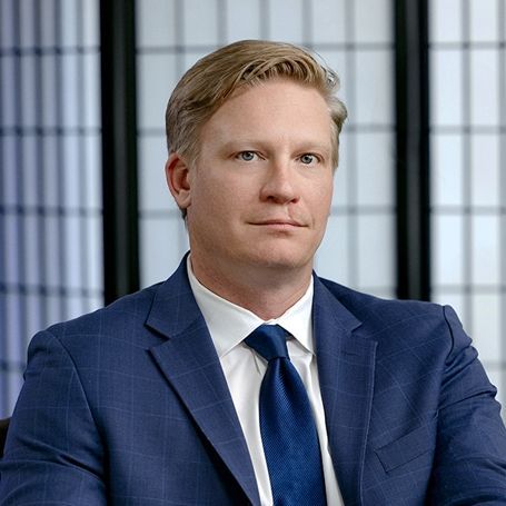A person with light hair wearing a blue plaid suit and blue tie, posed in front of a white grid screen.