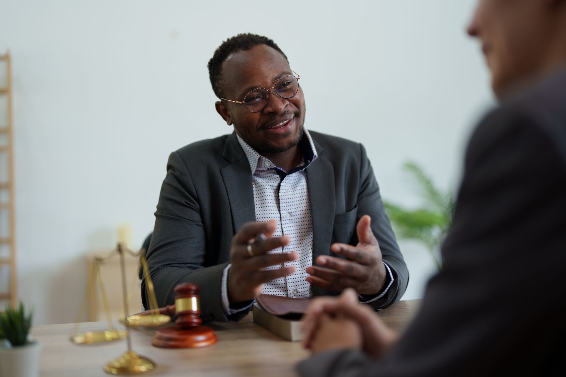 A professional sitting at a desk with a wooden gavel and scales of justice, gesturing while speaking to a client.