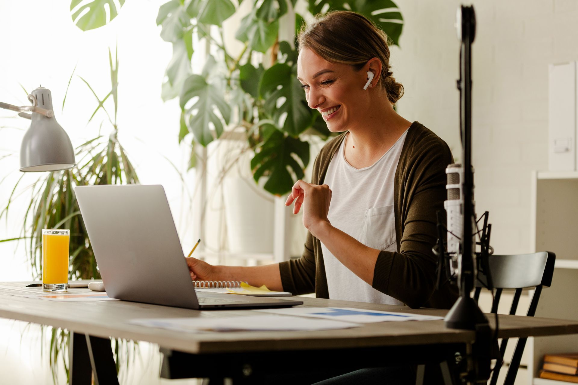 A smiling person sits at a desk with a laptop and a glass of orange juice, working in a room with a large indoor plant.