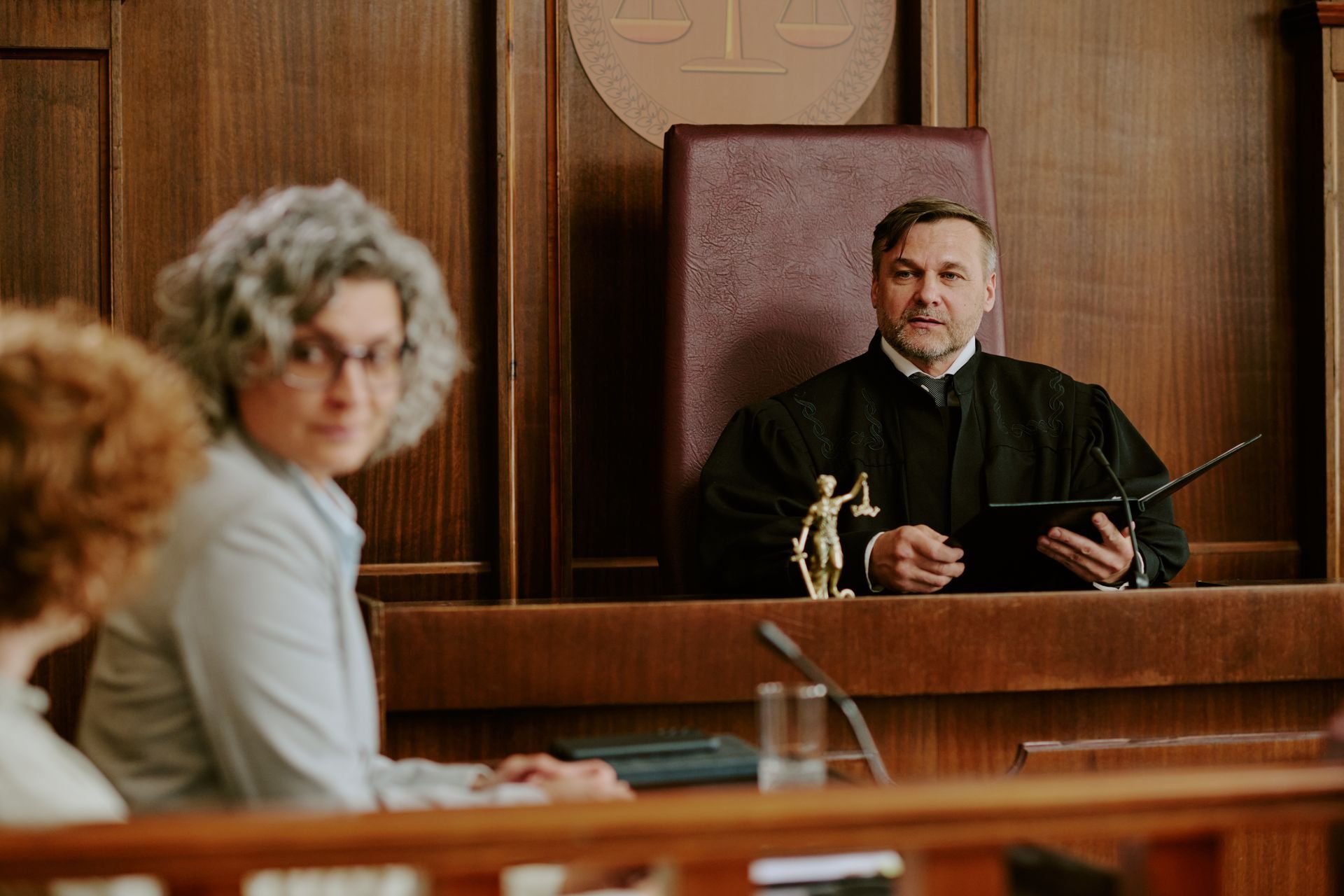 A judge in a black robe sits at a wooden bench in a courtroom, looking toward people seated in the foreground.