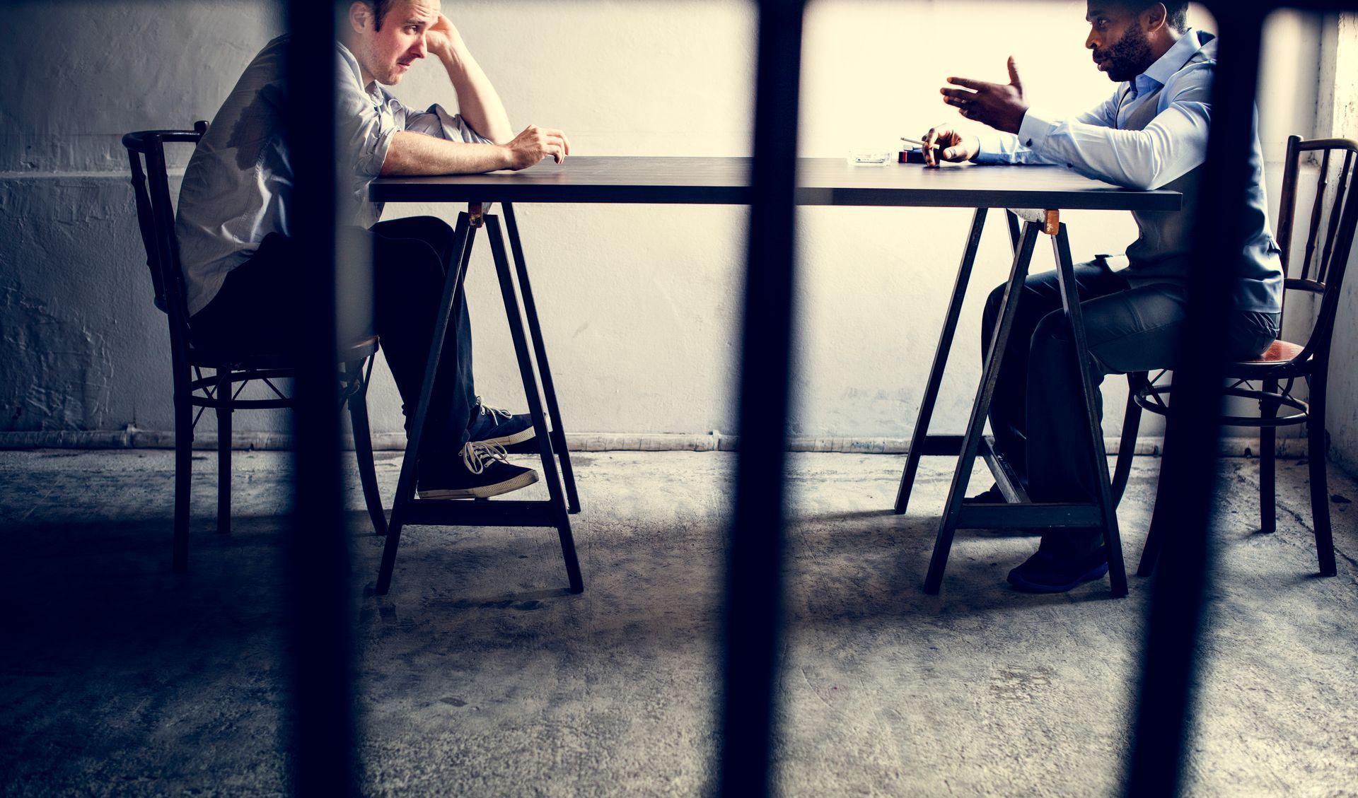 Two people sit at a table facing each other in a room viewed through vertical metal bars, resembling a jail visit.