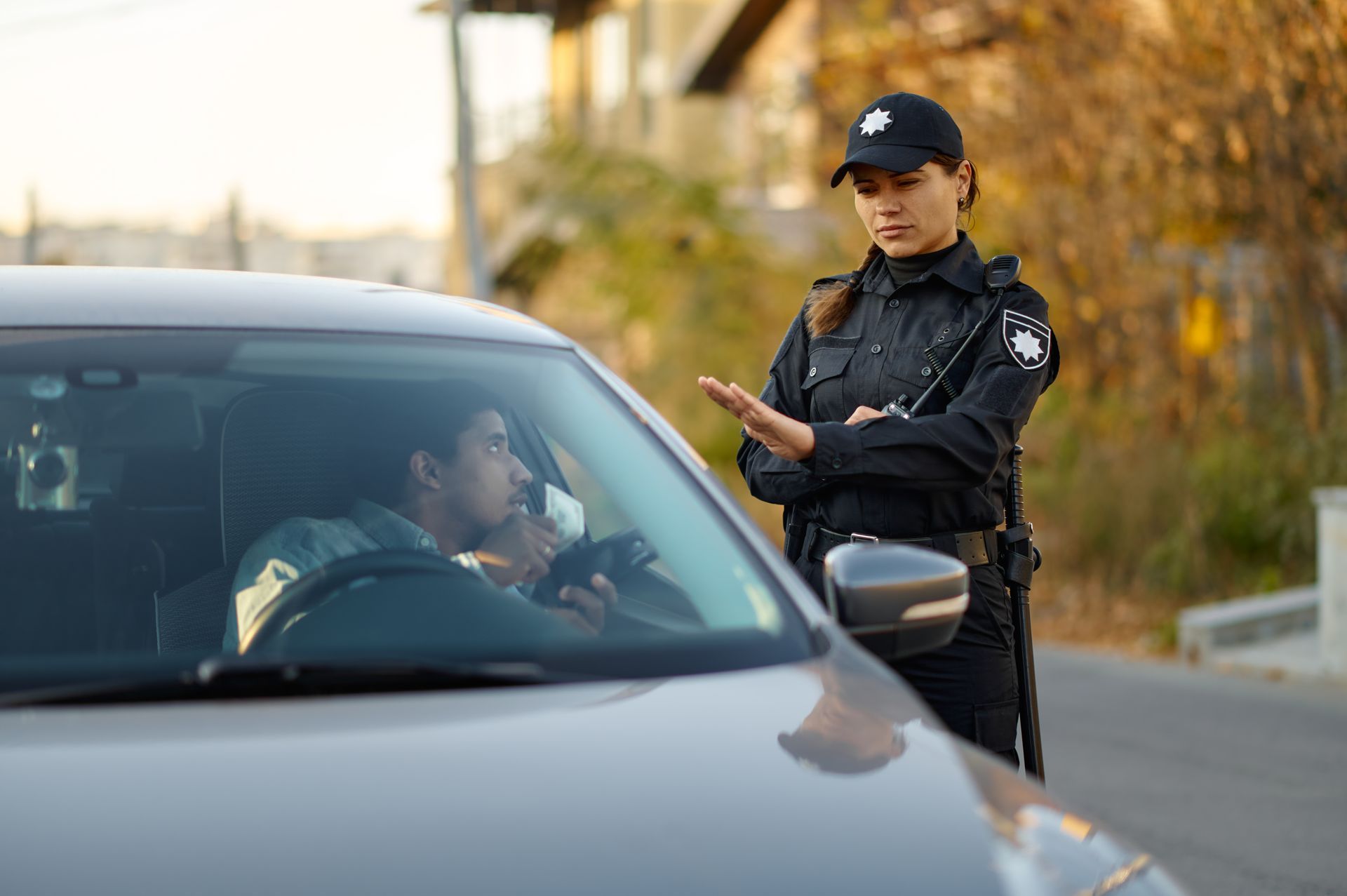 A police officer stands outside a car, gesturing to a driver who is using a breathalyzer in the driver's seat.