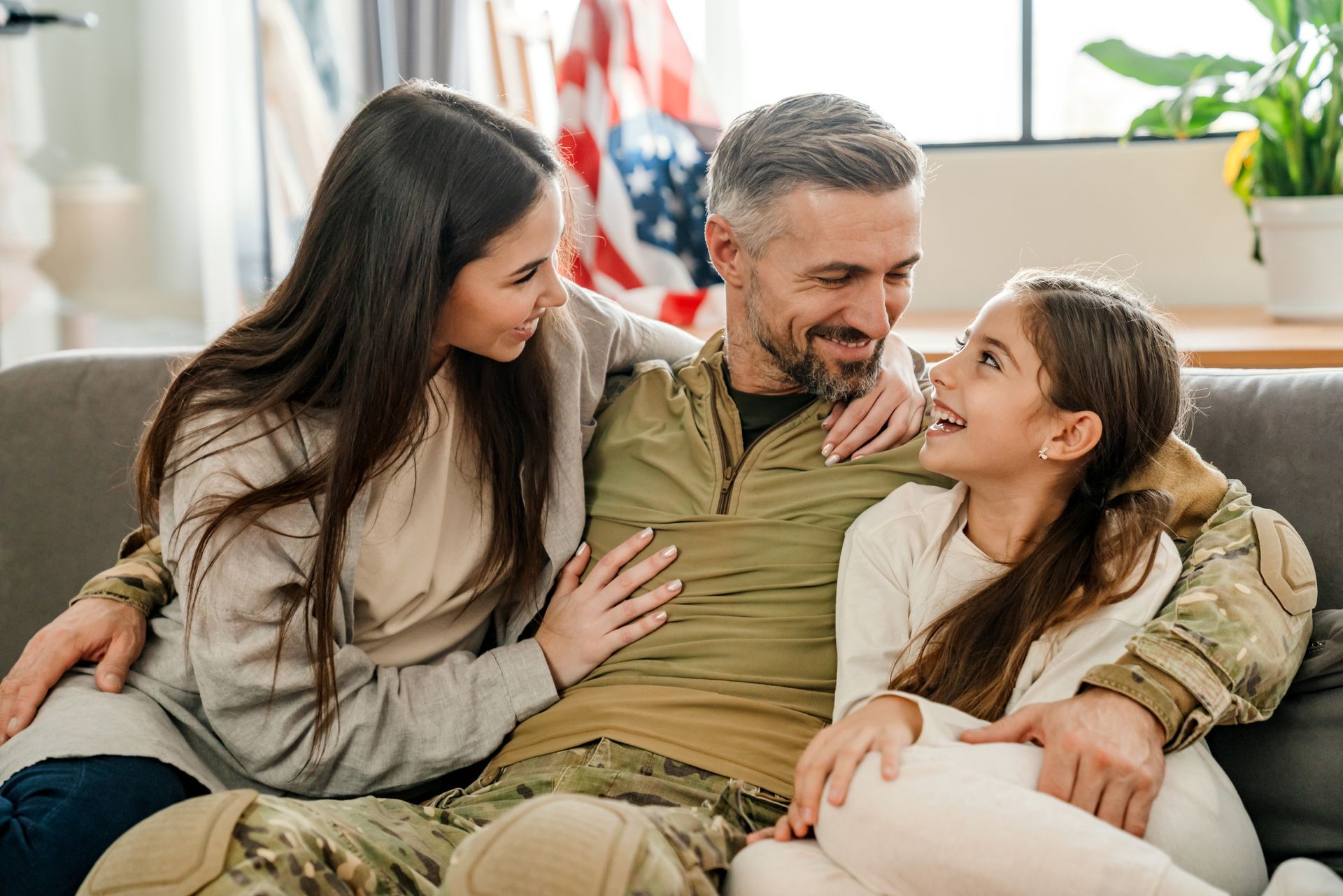 A family in military uniform sitting on a sofa, embracing and smiling in a home setting with an American flag.