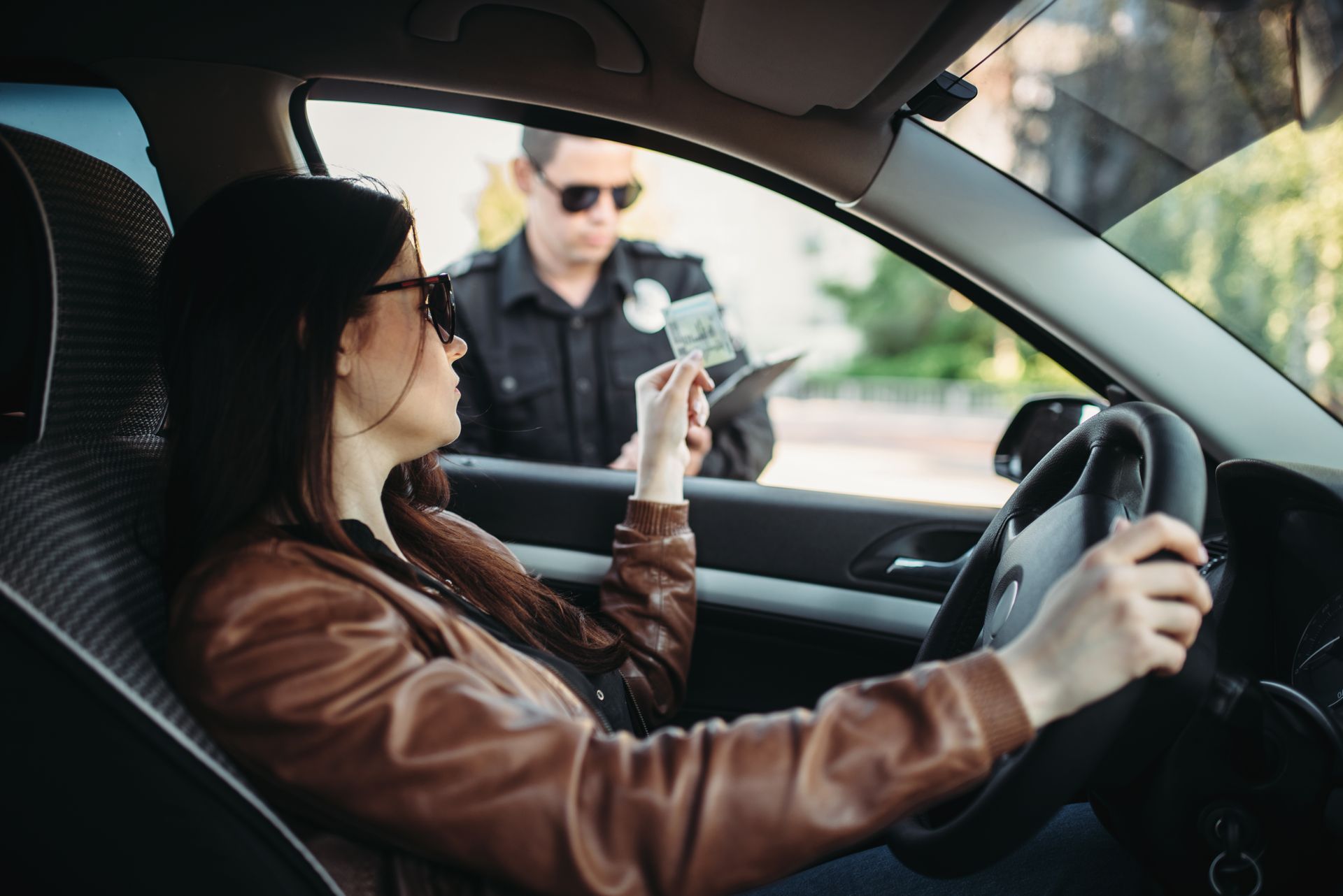 A driver in a brown jacket hands cash through a car window to a police officer wearing sunglasses.