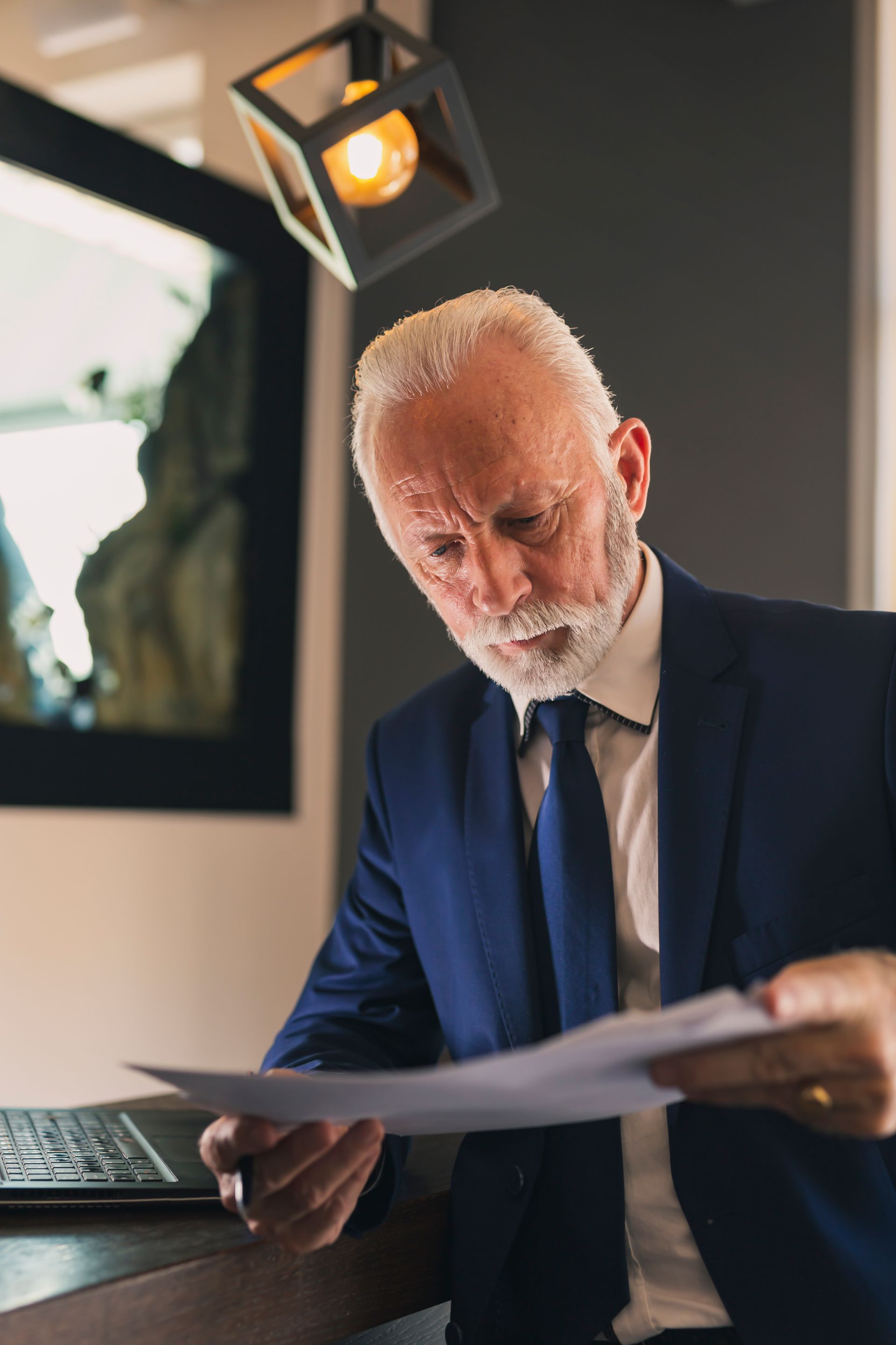 A focused person in a navy suit and tie reviews documents at a desk under a hanging geometric light fixture.