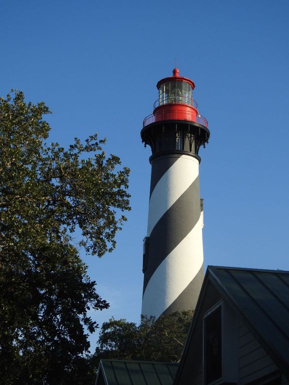 St. Augustine Lighthouse with its iconic black and white spiral stripes stands against a clear blue sky behind some trees.