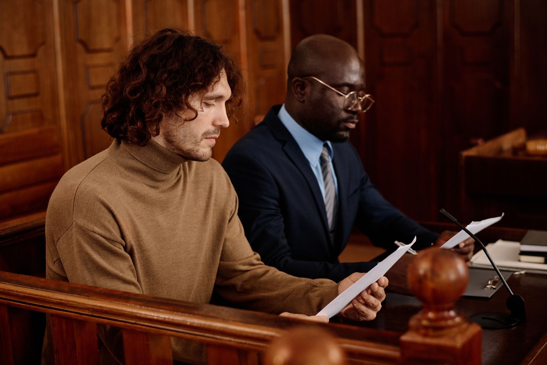 A person in a brown turtleneck and a lawyer in a suit review legal documents in a wood-paneled courtroom.