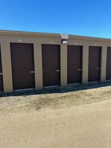 Row of brown storage unit doors with beige walls, gravel ground, and a blue sky.