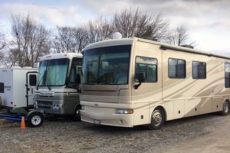 RVs parked outside on a gravel lot. Beige and white motorhomes with a small trailer in the background. Overcast sky.