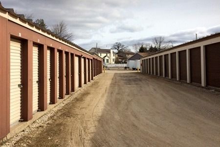 Rows of storage units with brown doors and tan frames, gravel driveway, cloudy sky.