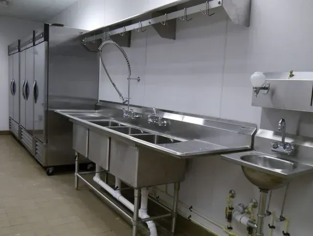 Commercial kitchen with stainless steel appliances: refrigerator, sinks, and hanging racks.