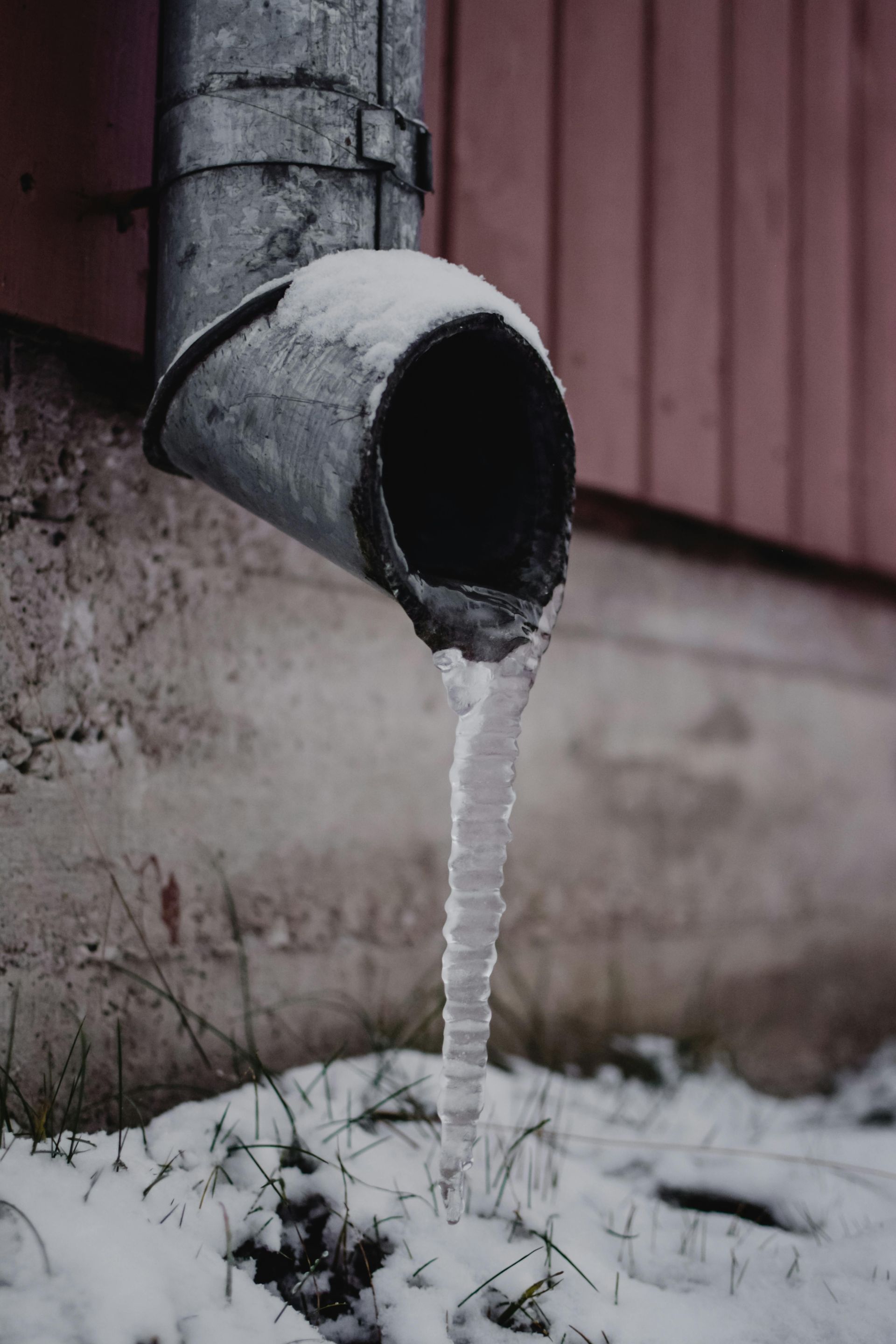 Icicle formed from a metal downspout, with snow on the ground and a red wooden wall in the background.