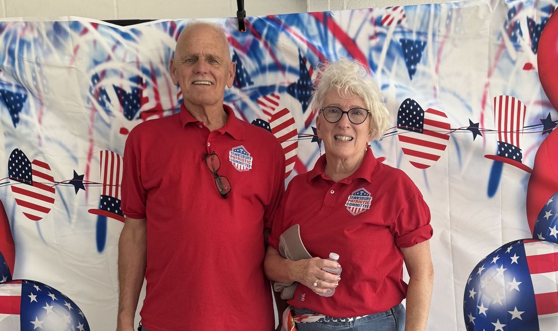 Two smiling people wearing red shirts with USA-themed buttons stand in front of a patriotic backdrop with balloons.