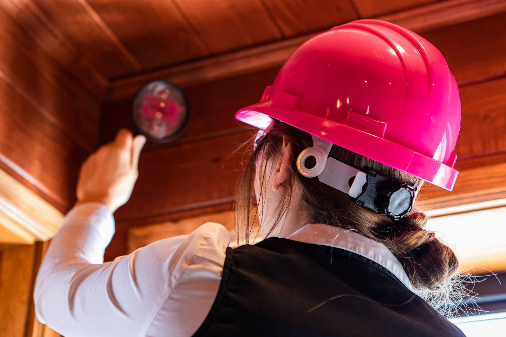 Person in a pink hard hat inspecting an object on a wooden wall.
