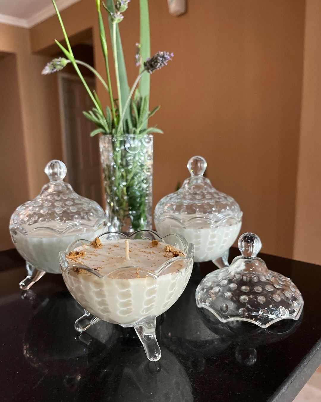 Tres bomboneras de cristal sobre una mesa negra con un jarrón de flores al fondo.