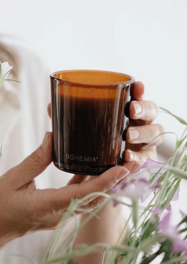 Una mano sostiene una taza de vidrio marrón cerca de plantas de color verde suave sobre un fondo claro.