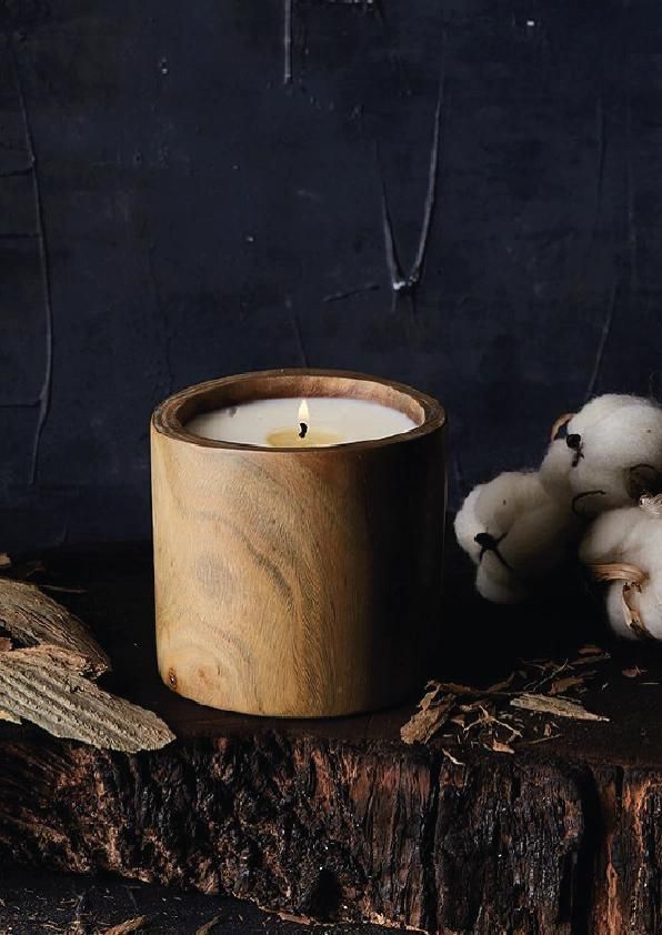 Vela beige encendida en un portavelas de madera junto a tallos de algodón sobre una superficie rústica de madera oscura.
