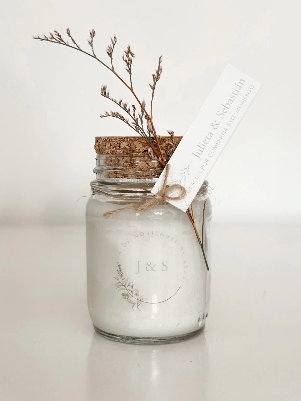 Vela en tarro de cristal con flores secas y etiqueta blanca sobre fondo claro.
