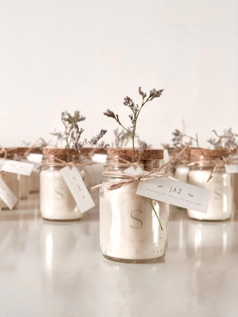 Pequeños frascos de vidrio con velas blancas, cordel y flores secas sobre un fondo claro.