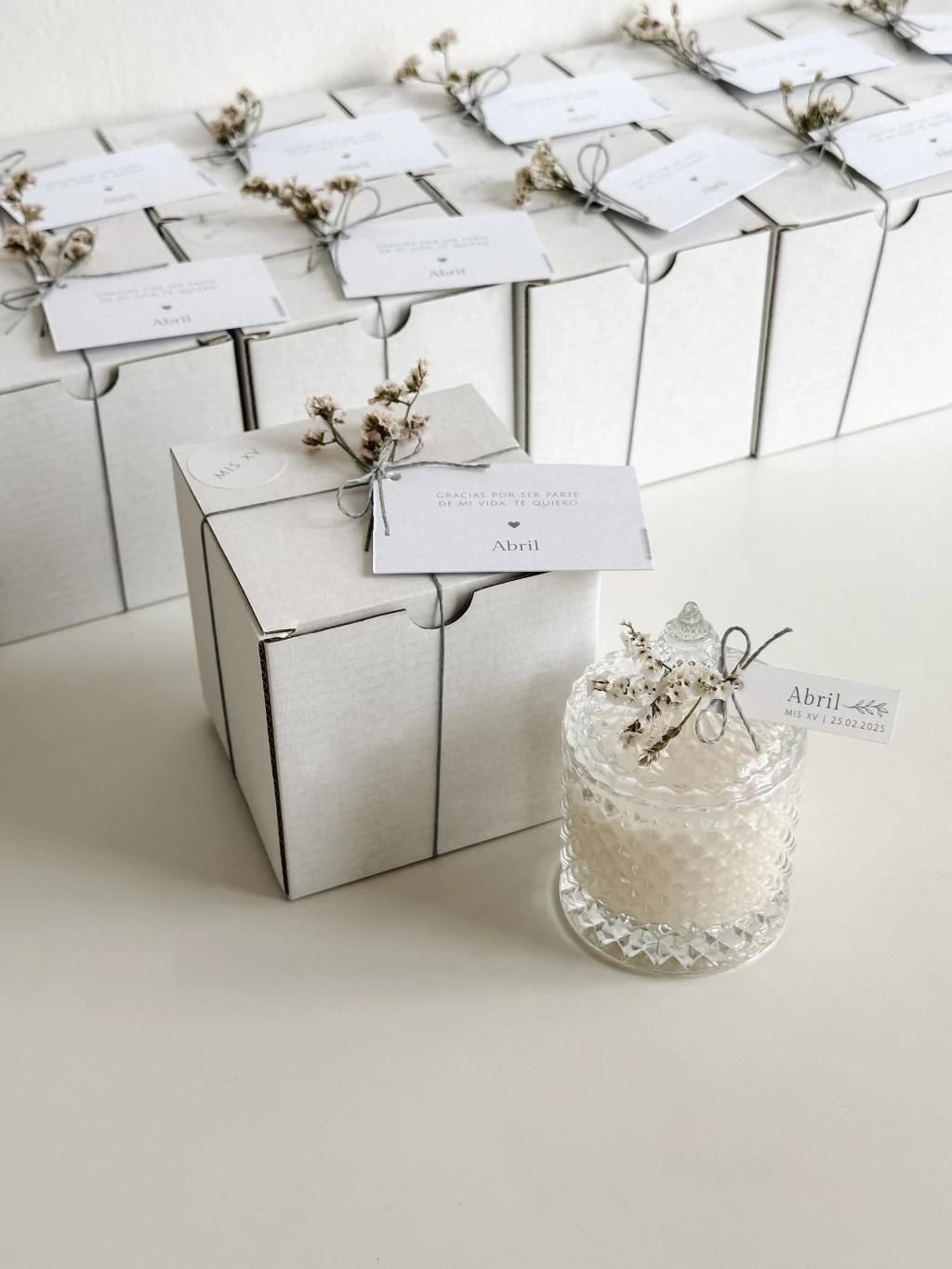 Cajas de regalo con cintas plateadas y una pequeña vela blanca sobre una superficie blanca.