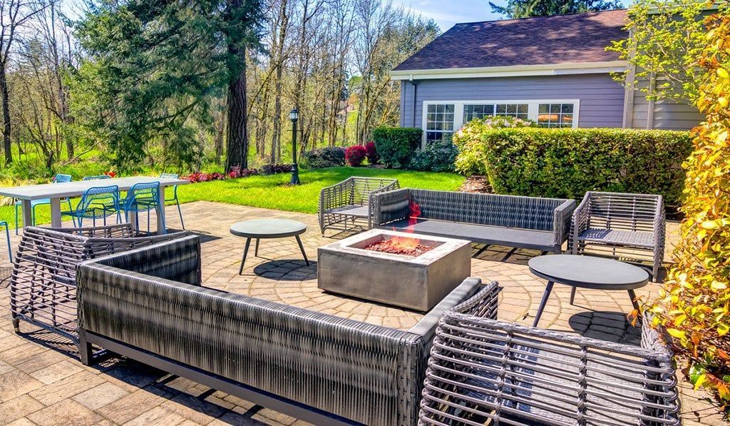Patio with wicker seating, fire pit, and small tables, in front of a blue house and green trees.