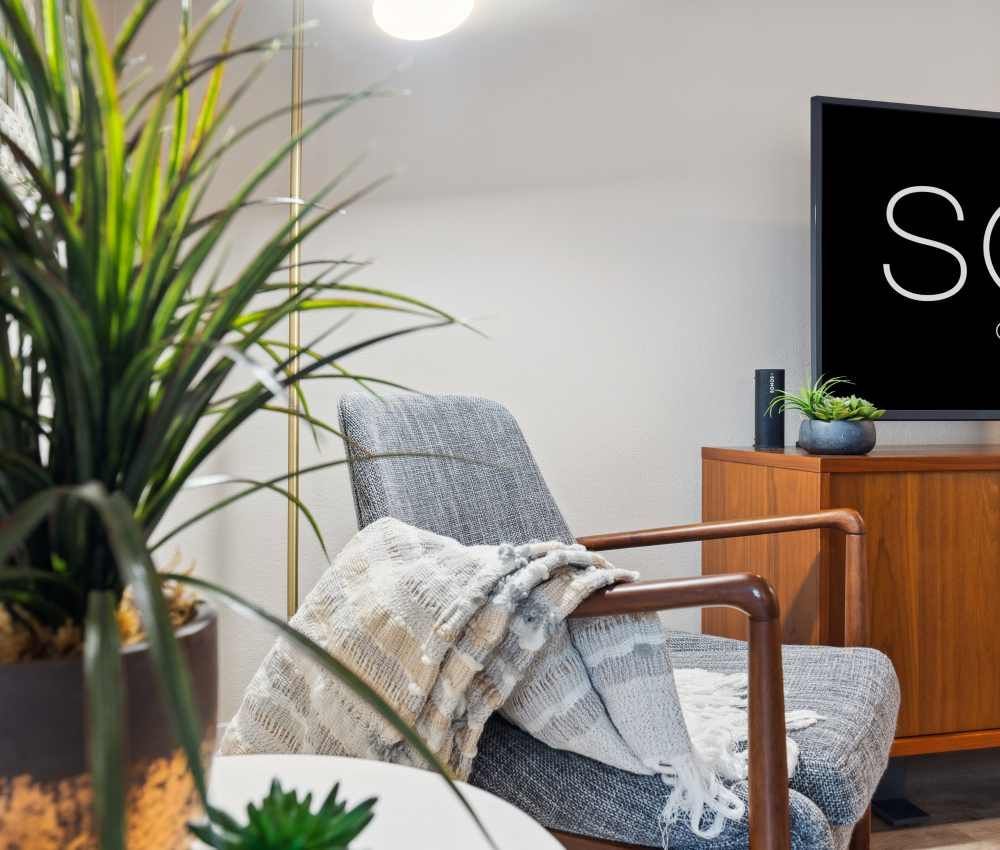Modern living room with a gray upholstered chair, cozy throw, and a TV on a wooden console at Sofi at Murrayhill, offers apartments near Murrayhill Marketplace.