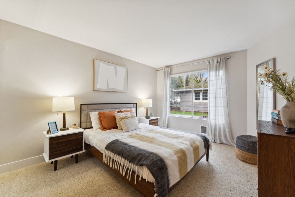 Bedroom with a bed, two nightstands, lamps, dresser, and a large window with sheer curtains at Sofi at Murrayhill, offers apartments near Murrayhill Town Center.