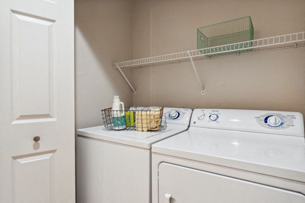 Laundry room with a washing machine and dryer, white door, and wire shelf with items at Sofi at Murrayhill, offers apartments near Murrayhill Town Center.