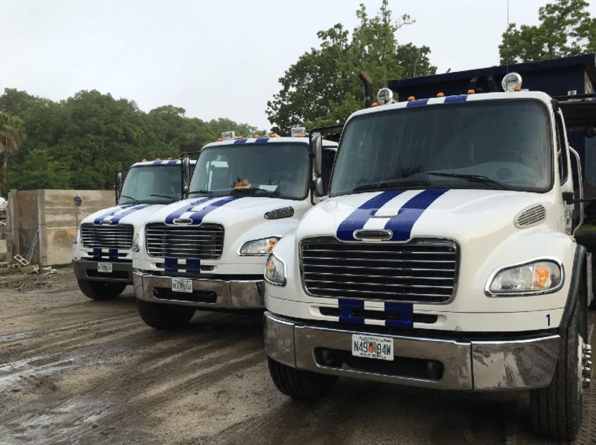 A row of white trucks with blue stripes on the front are parked in a dirt lot.