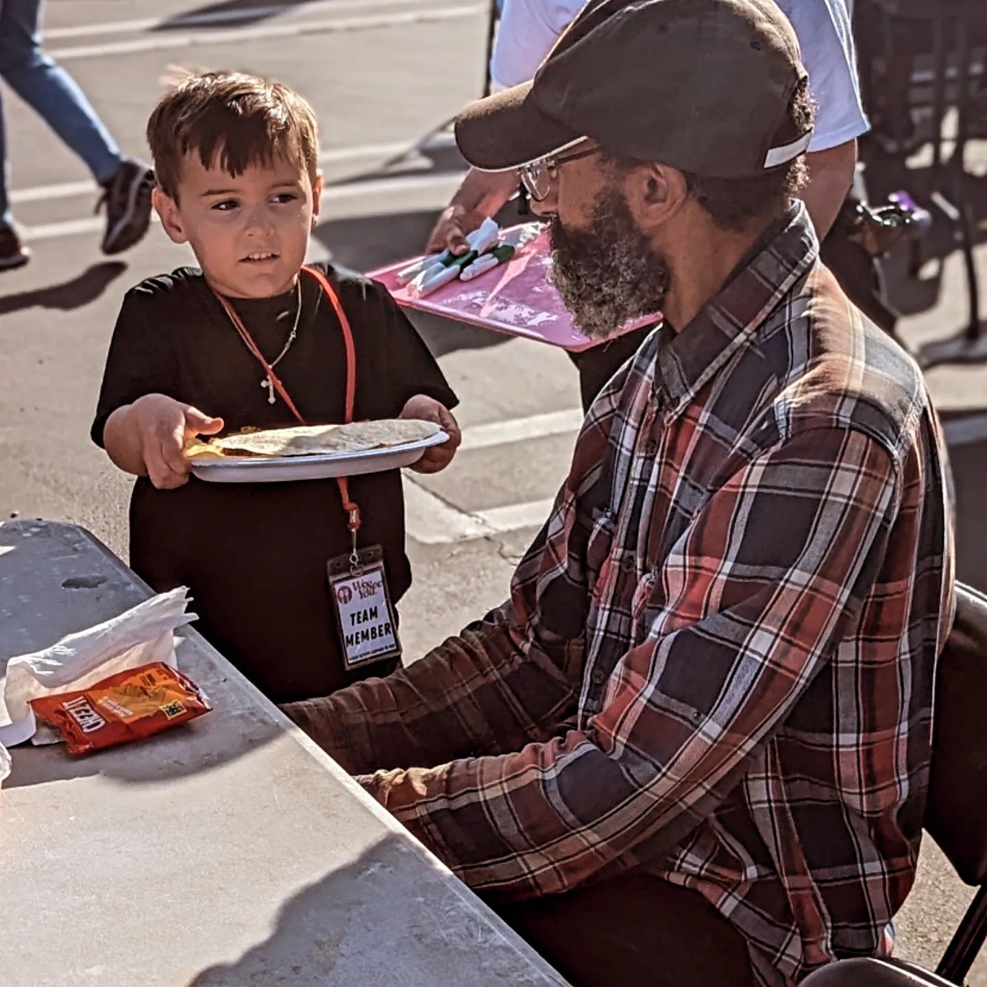 A man in a plaid shirt is sitting at a table with a little boy holding a tray of food