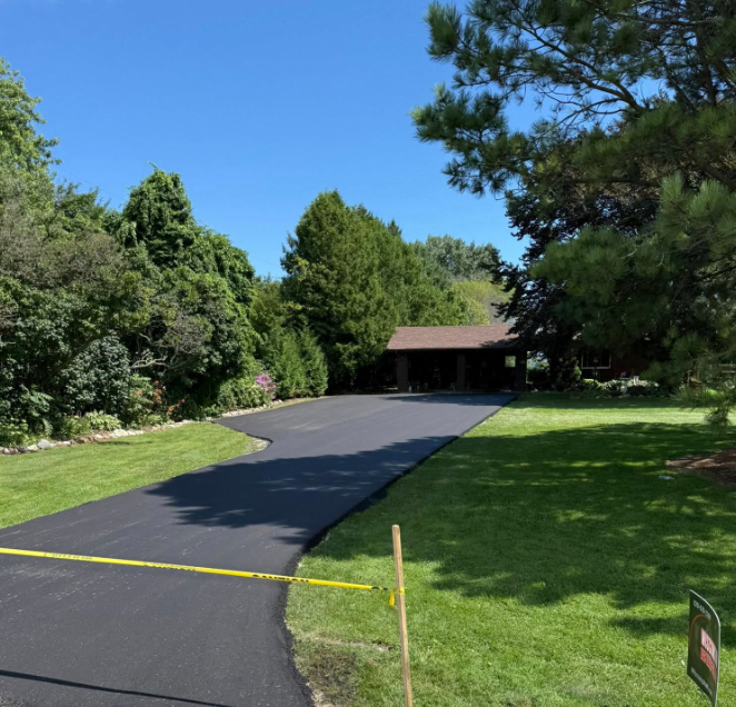 Newly paved asphalt driveway leading to a brown house with trees and green lawn under a blue sky.