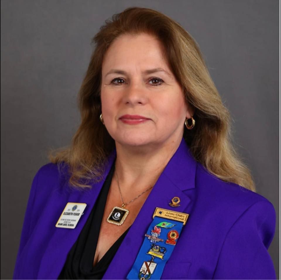 Woman with blond hair, wearing a purple blazer and ribbon. She's looking at the camera, possibly at an event.