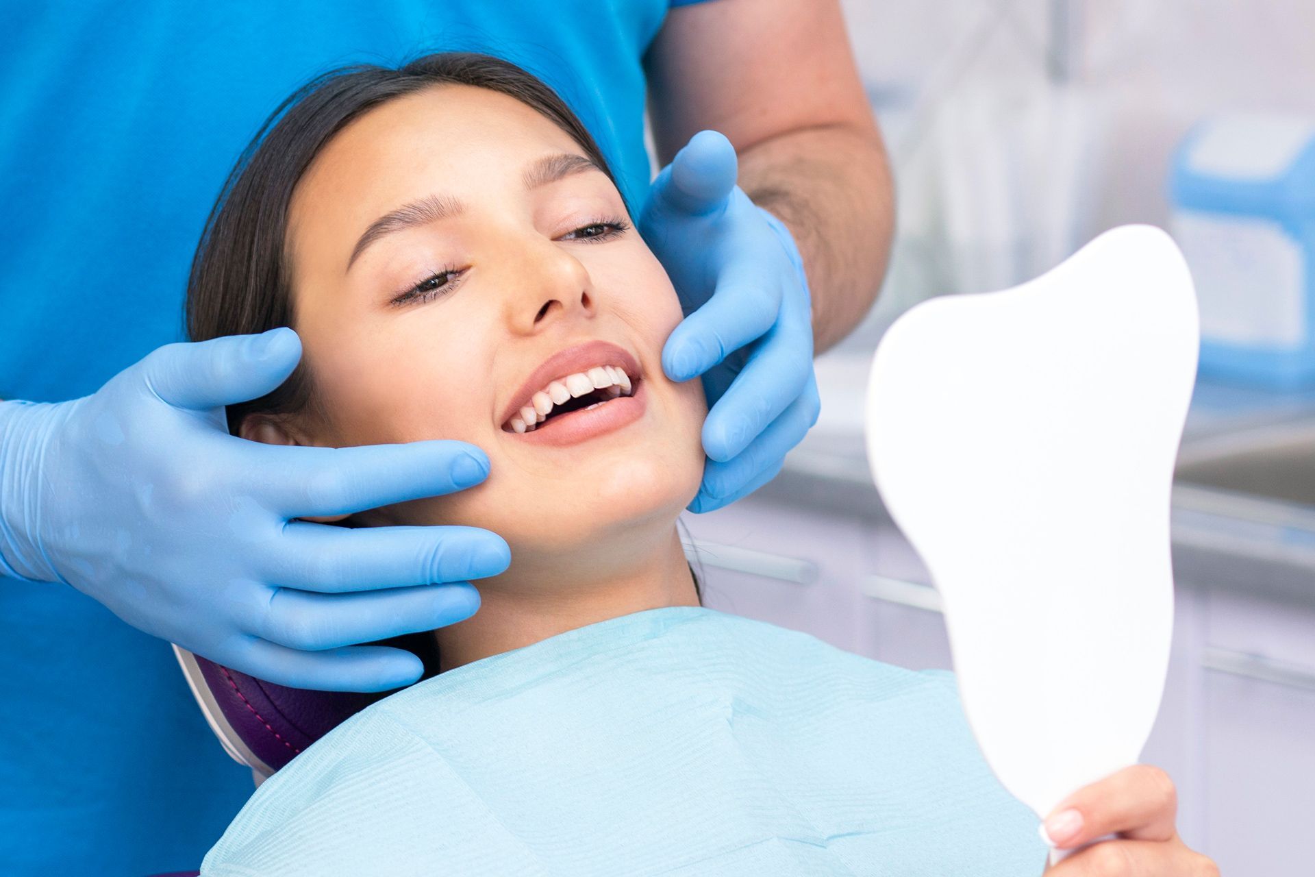 A woman is sitting in a dental chair looking at her teeth in a mirror.