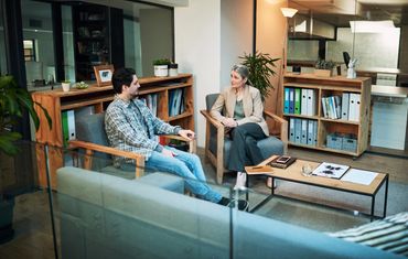 Two people sit in armchairs in a modern office, having a conversation across a coffee table with documents.