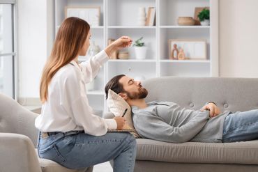 A therapist in a white shirt holds a pendulum over a person lying on a couch during a hypnotherapy session.