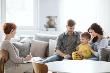 A therapist in a bright office takes notes as a family with a young child playing with a yellow die sits on a sofa.