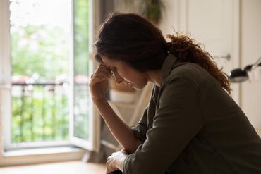 A person sits in profile, resting their forehead on their hand, looking down with a somber expression in a bright room.