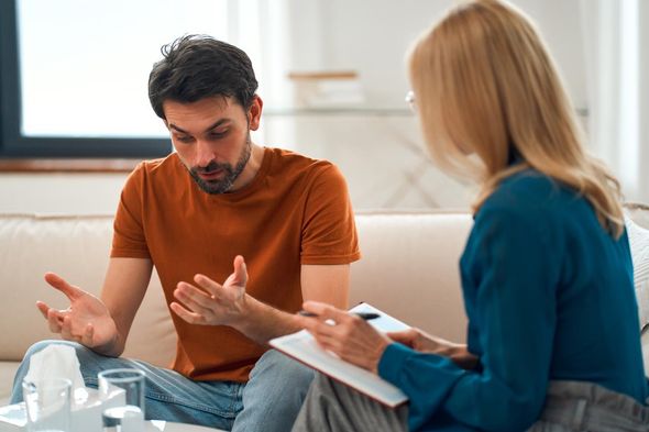 A person in an orange t-shirt sits on a couch gesturing while talking to a therapist who is taking notes.