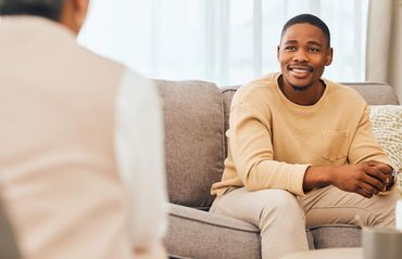 A person in a beige sweater sits on a grey sofa, smiling while speaking with someone off-camera in a bright room.