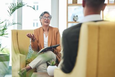A professional in a tan blazer sits in a yellow chair, gesturing while speaking to someone seated across from them.