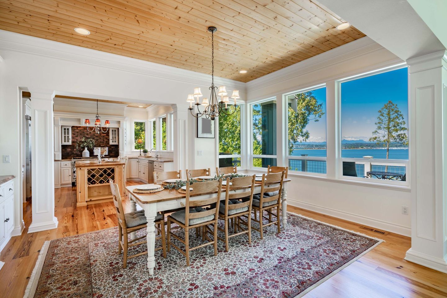 Dining room with long table, chandelier, large windows with water view, wood ceiling and flooring.