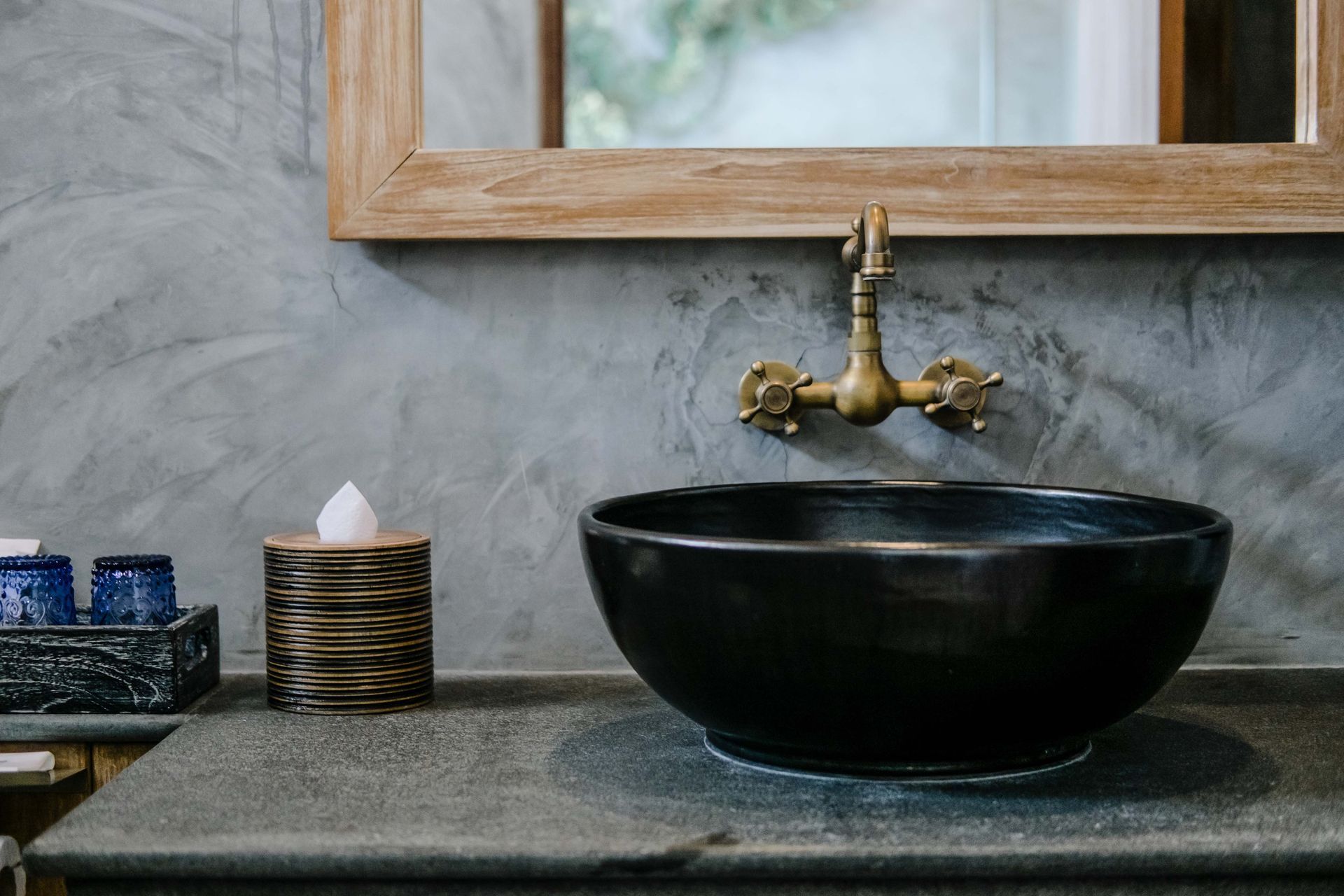 Black bowl sink, brass faucet, wooden mirror frame, on gray countertop.