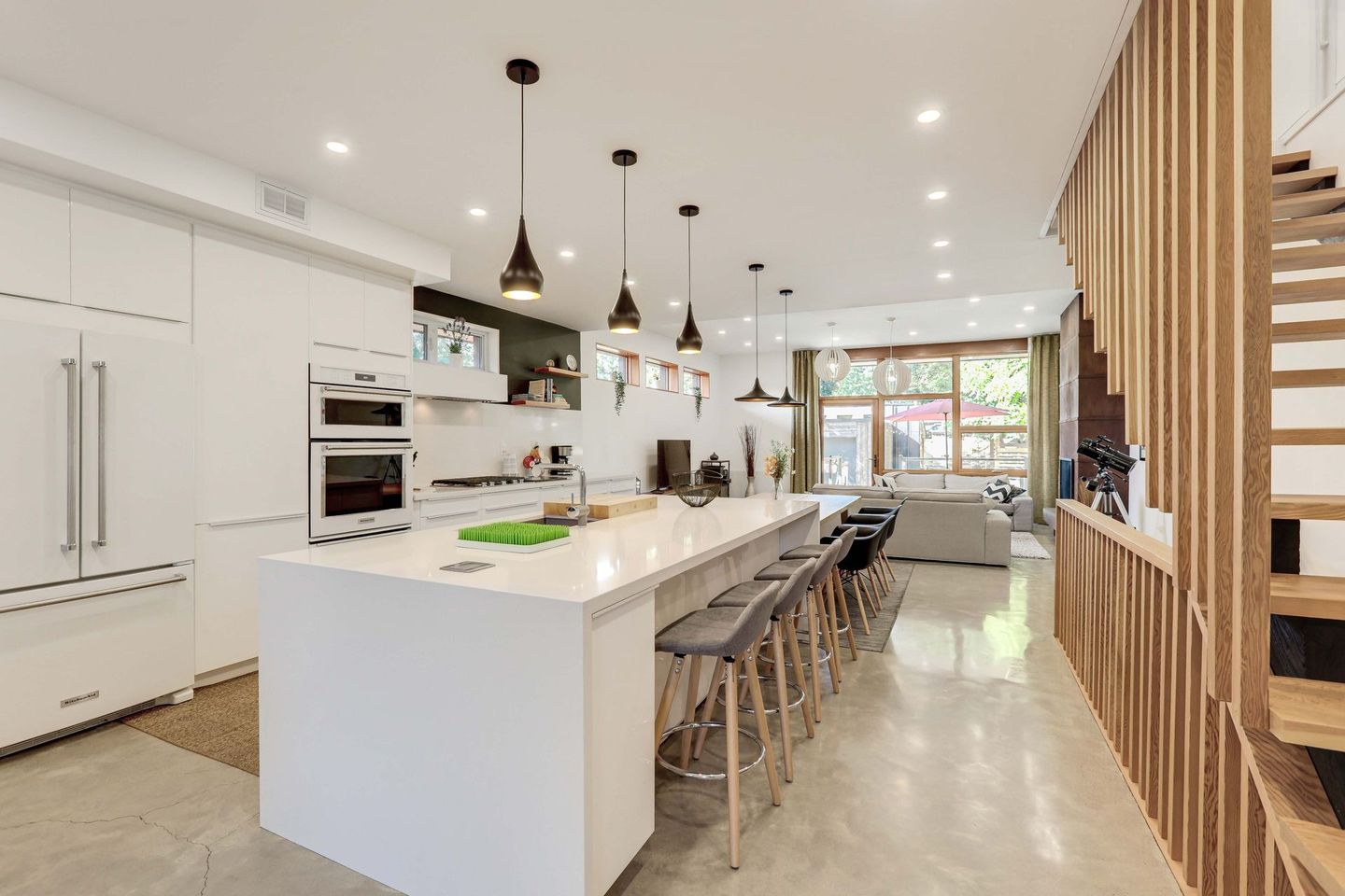 Modern white kitchen with island seating, leading to a living area with a staircase.