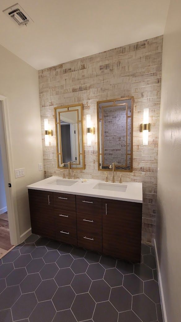 Interior of a modern bathroom with a double vanity featuring two vessel sinks and chrome faucets. The vanity is topped with a white quartz countertop and framed by a large mirror with sconces on either side. Light gray ceramic tile covers the floor 