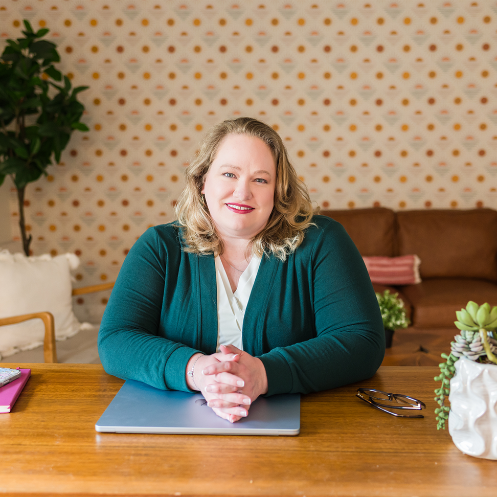 Woman at desk in green sweater smiles; laptop, notebook, and succulent present.