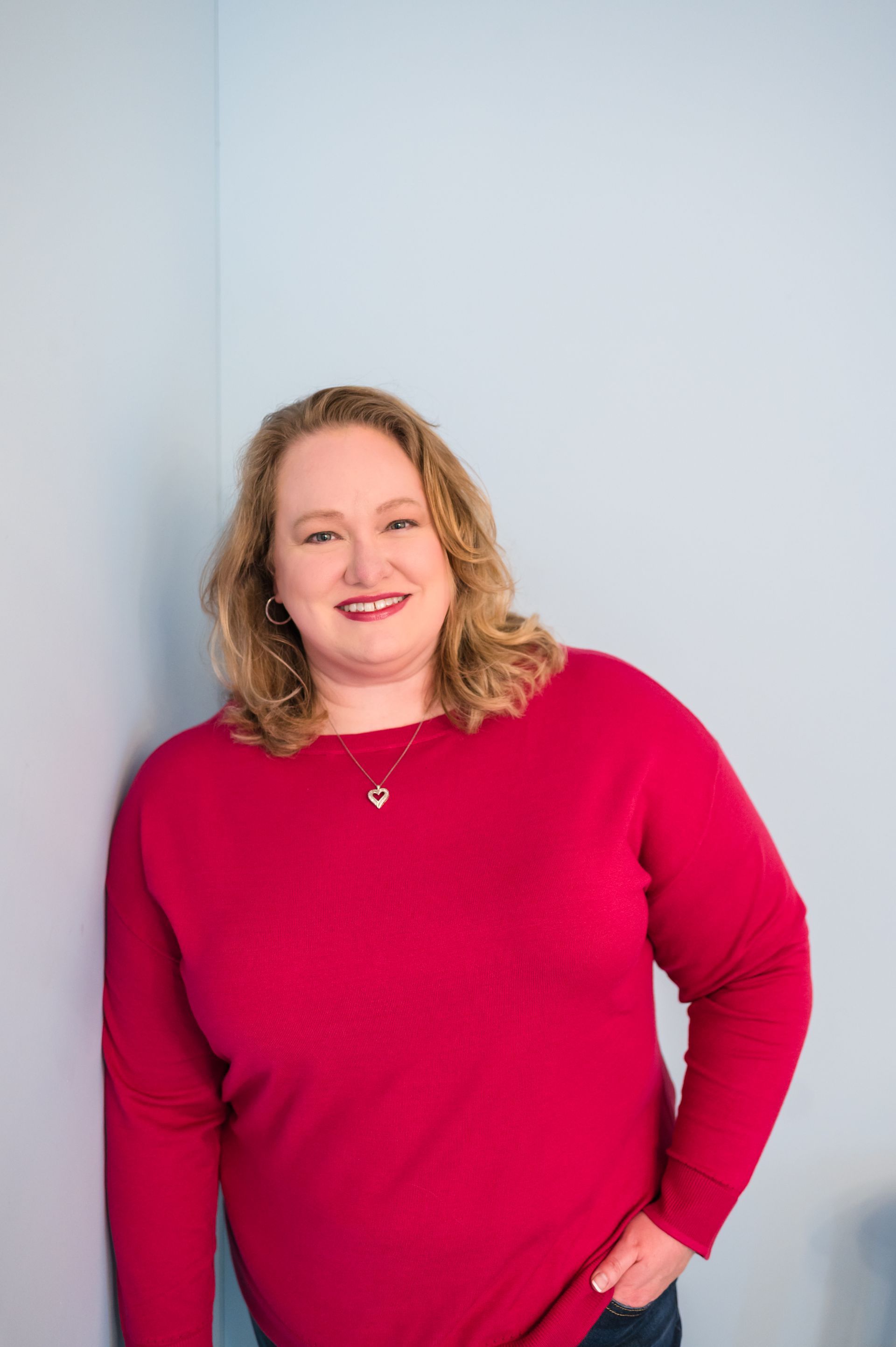 Woman in a red sweater smiles, leaning against a light blue wall.