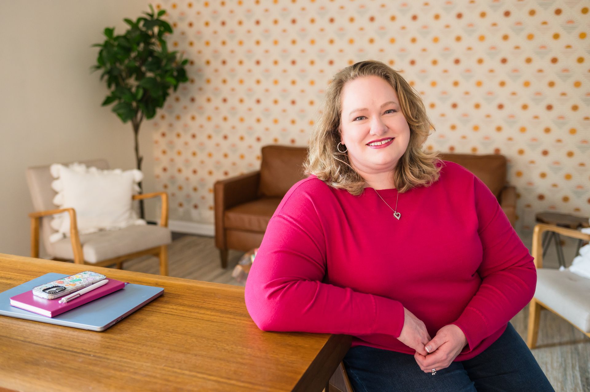 Woman in pink top smiles, leaning on wooden desk with laptop. Interior setting with tan wallpaper.