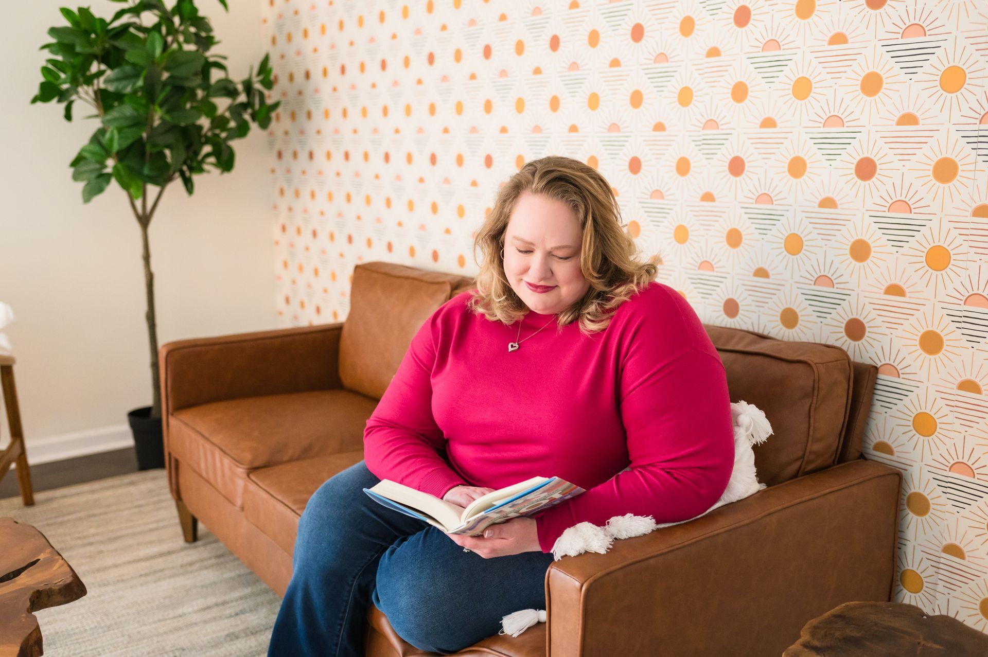 Woman reading a book on a brown sofa in a room with patterned wallpaper and a potted tree.
