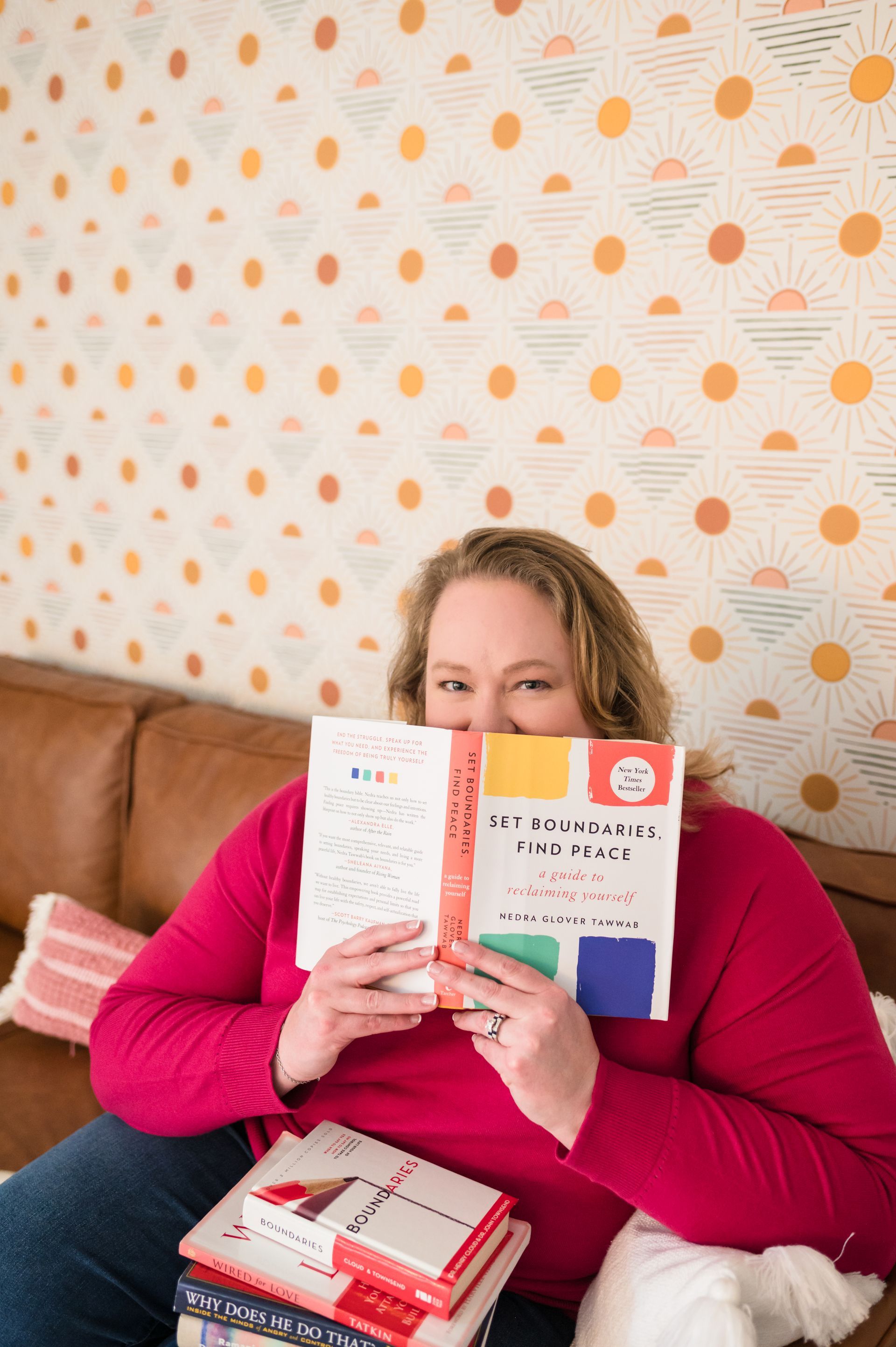 Woman holding open book, peeking over its pages, sitting on a brown couch. Colorful wallpaper in the background.